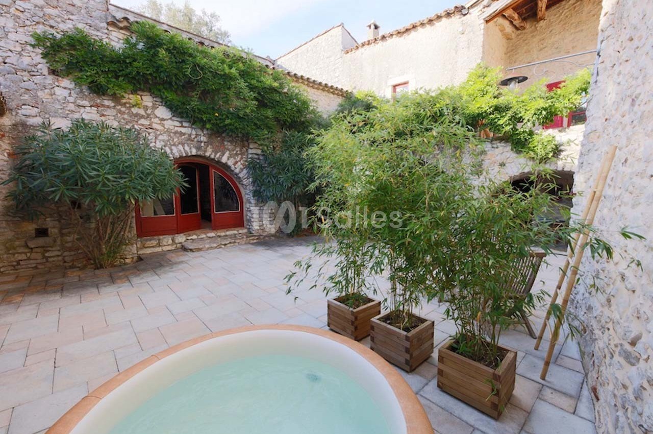 Cour intérieure en pierre avec jacuzzi, plantes en pots et façade de maison ancienne avec fenêtres et volets rouges.