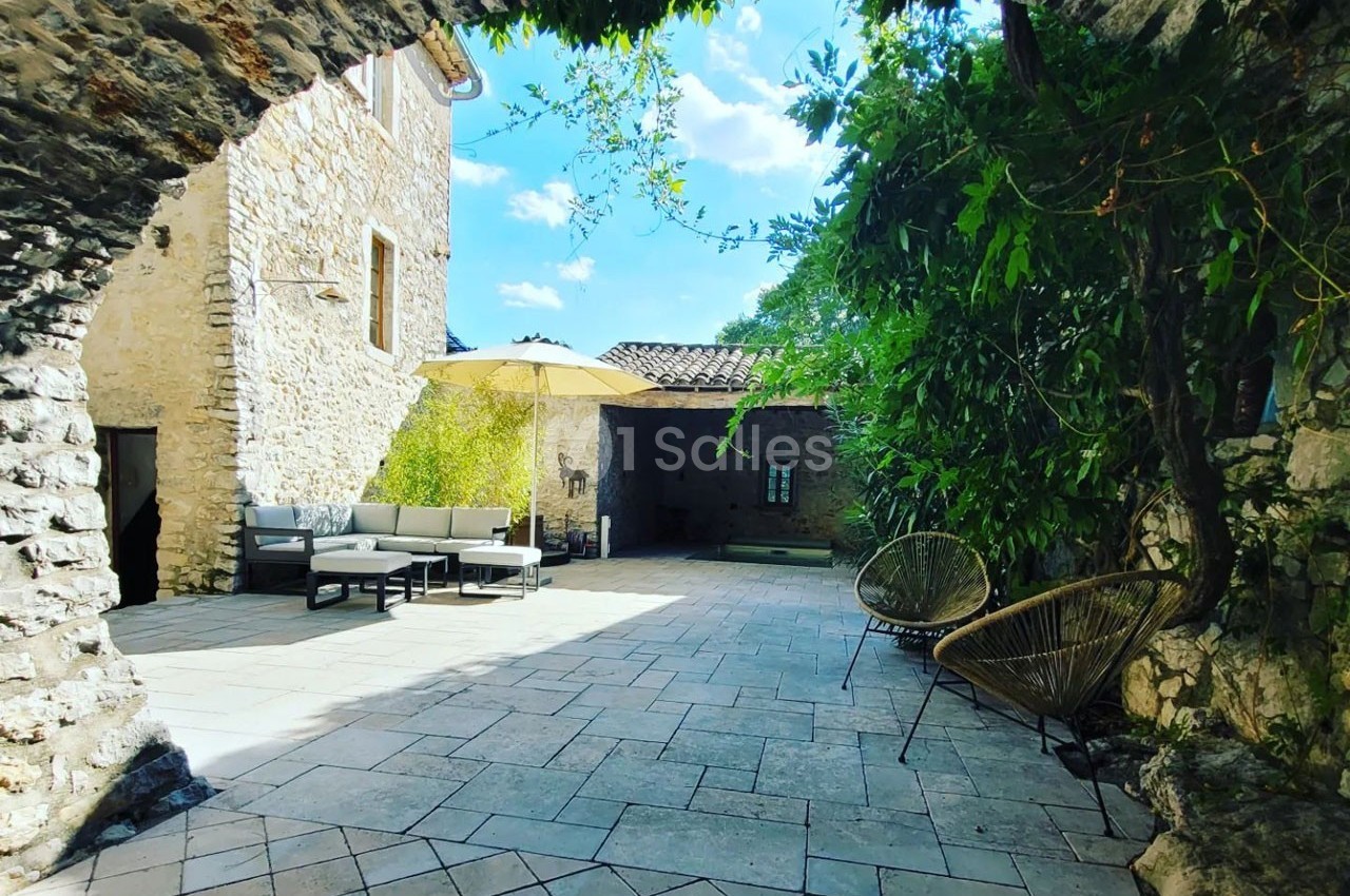 Cour intérieure pavée avec mobilier de jardin, parasol et végétation, entourée de murs en pierre sous un ciel dégagé.