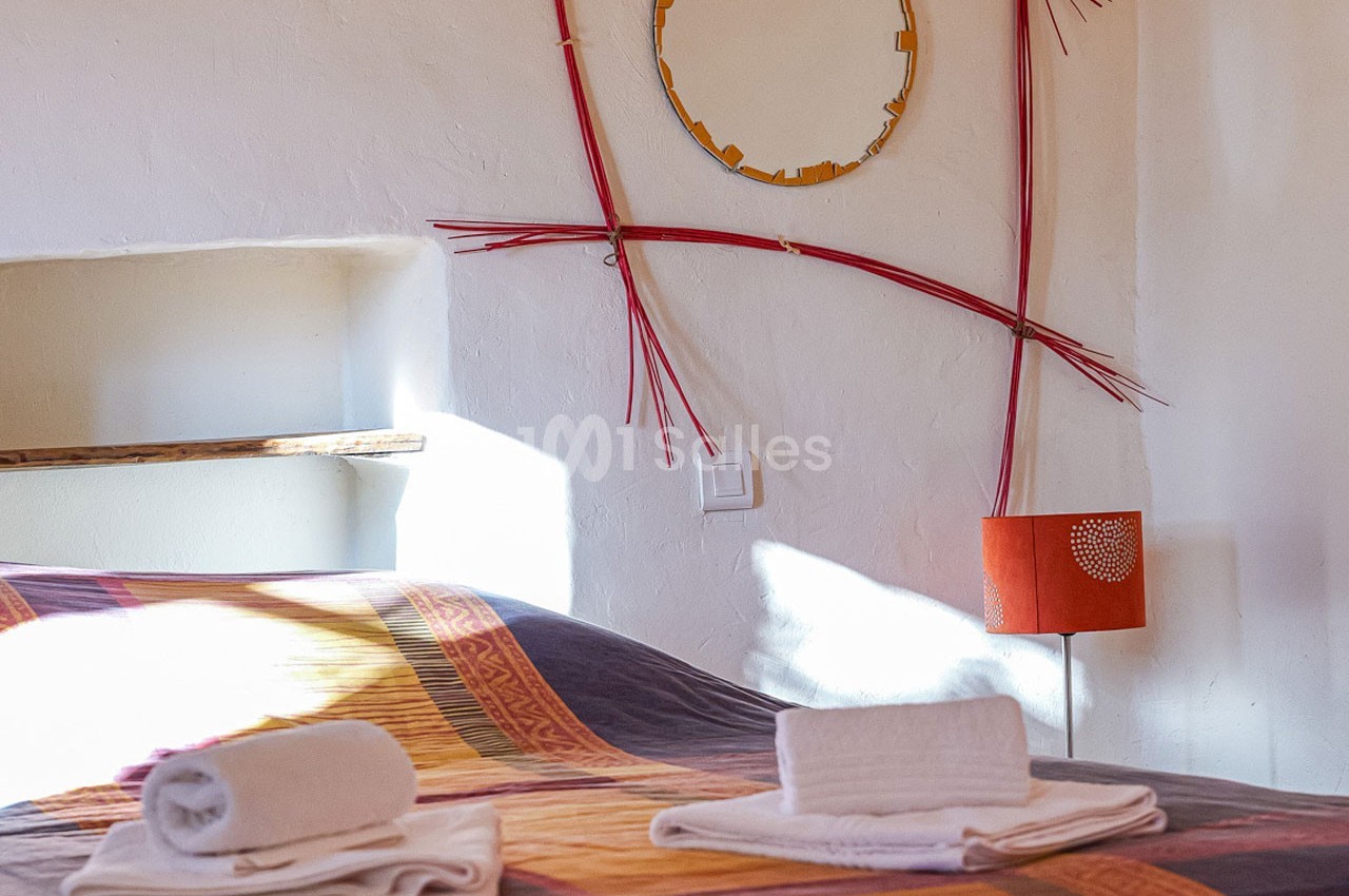 Lit avec couvre-lit coloré, serviettes pliées au premier plan, lampe orange et décoration murale en arrière-plan.