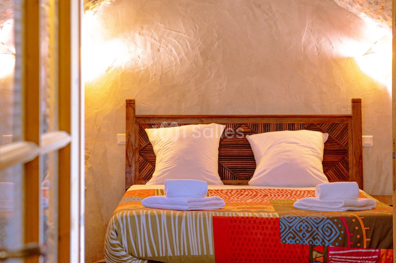 Lit double avec tête de lit en bois sculpté, draps colorés et deux serviettes pliées, dans une chambre aux murs en pierre.