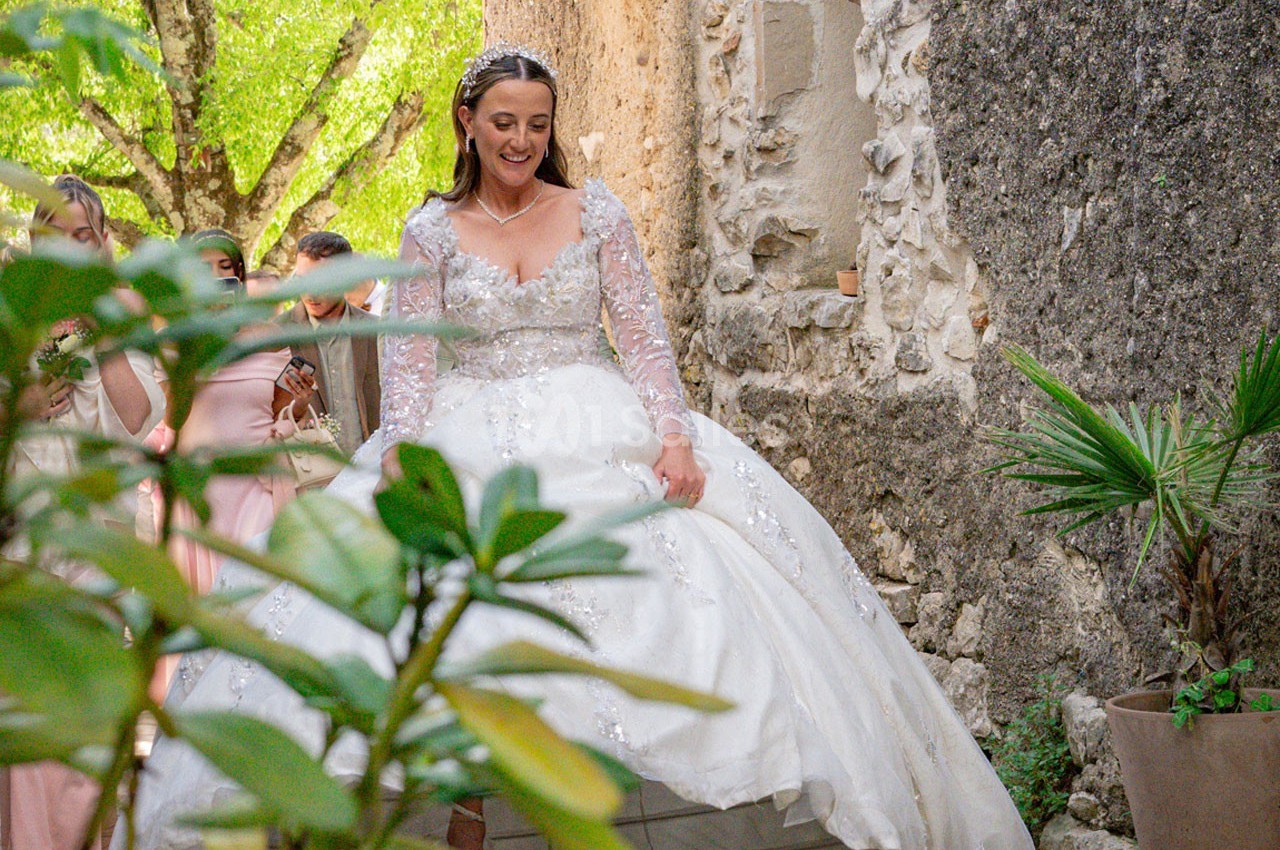 Une mariée en robe blanche ornée de dentelle descend des marches en pierre, entourée de verdure et d'invités.
