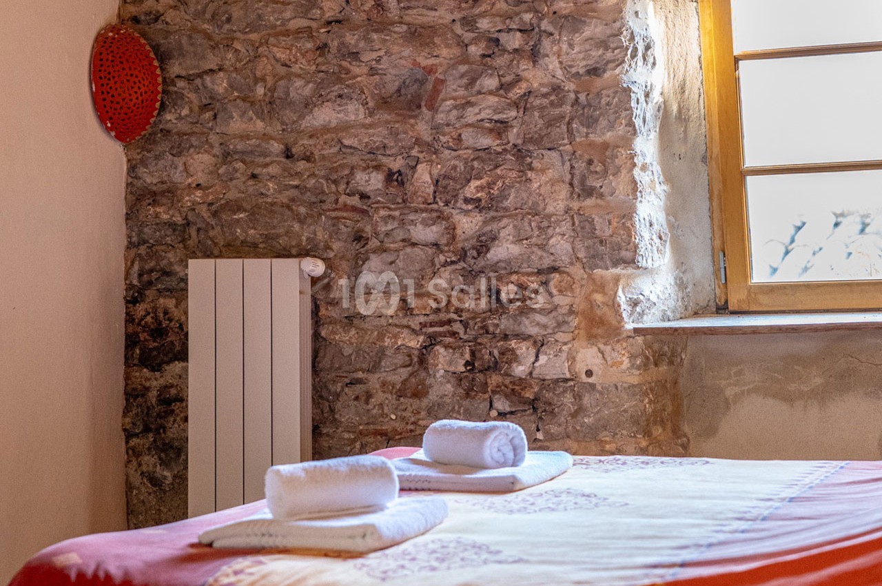 Lit recouvert d'un drap coloré avec deux serviettes roulées, dans une pièce aux murs en pierre et fenêtre en bois.