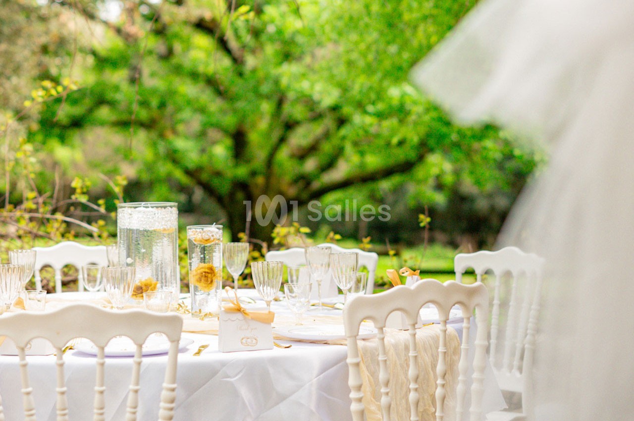 Table décorée avec des chaises blanches, nappes et verres élégants, installée en extérieur devant un arbre verdoyant.