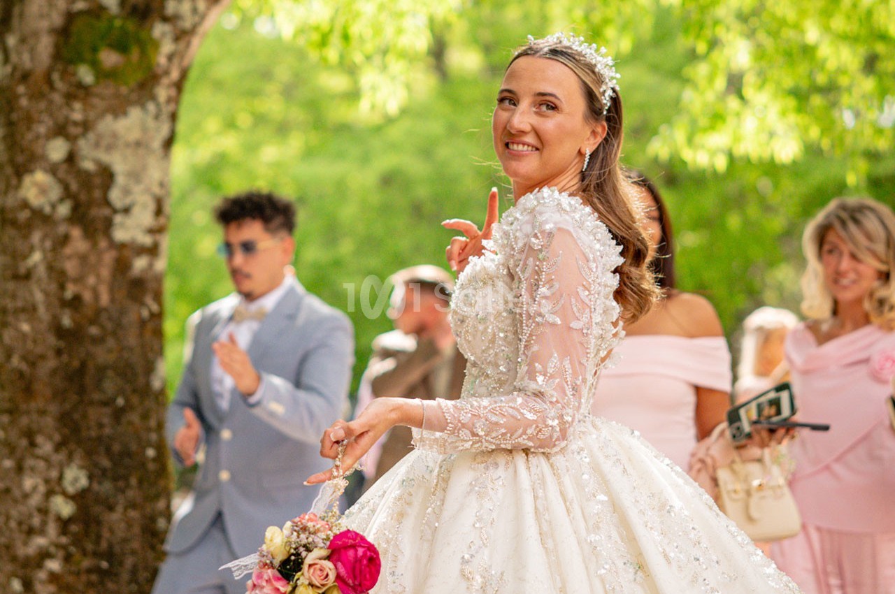 Une mariée souriante en robe blanche ornée de perles tient un bouquet, entourée d'invités dans un cadre extérieur verdoyant.
