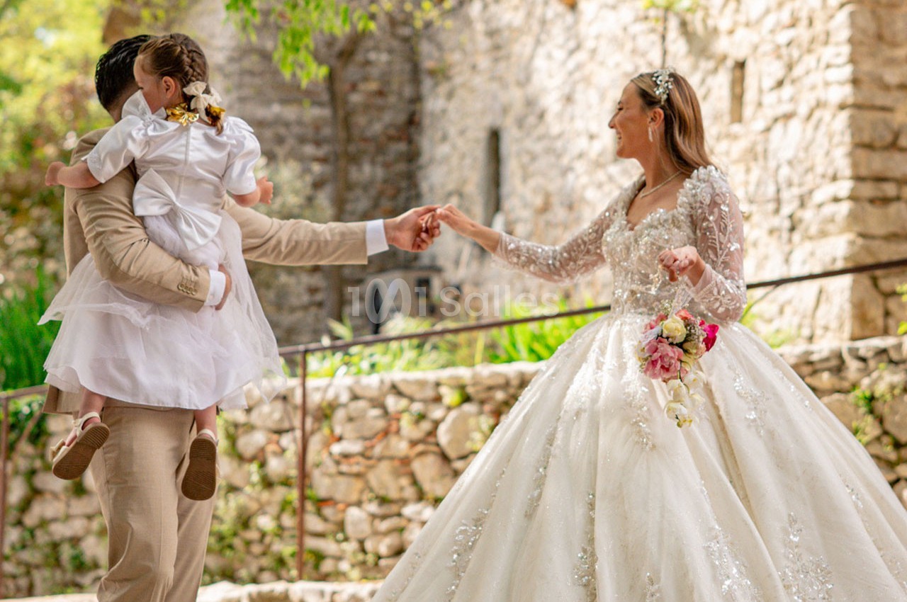 Une mariée en robe blanche tient la main d'un homme portant une enfant, devant un mur en pierre et des arbres.
