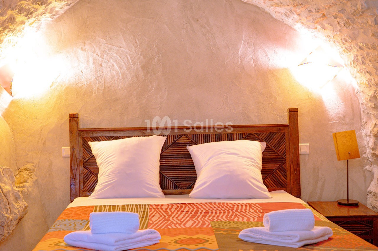Lit double avec tête de lit en bois sculpté, draps blancs et serviettes pliées, dans une chambre aux murs en pierre.