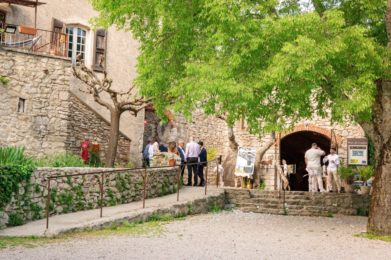 Des personnes discutent devant l'entrée voûtée d'un bâtiment en pierre, entouré d'arbres et d'un chemin pavé.