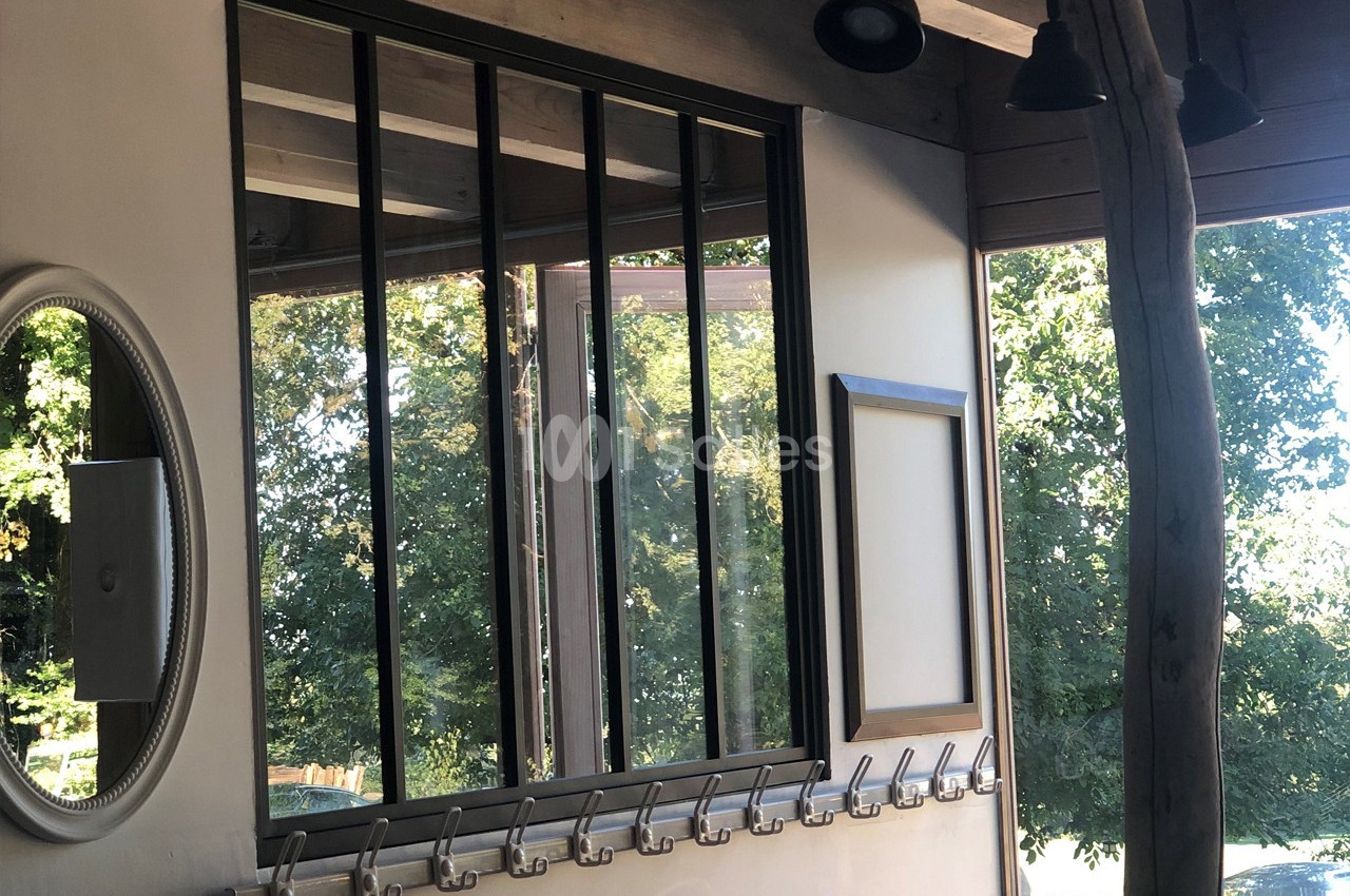 Mur intérieur avec verrière noire, miroir rond, patères métalliques et vue sur des arbres à travers une fenêtre.