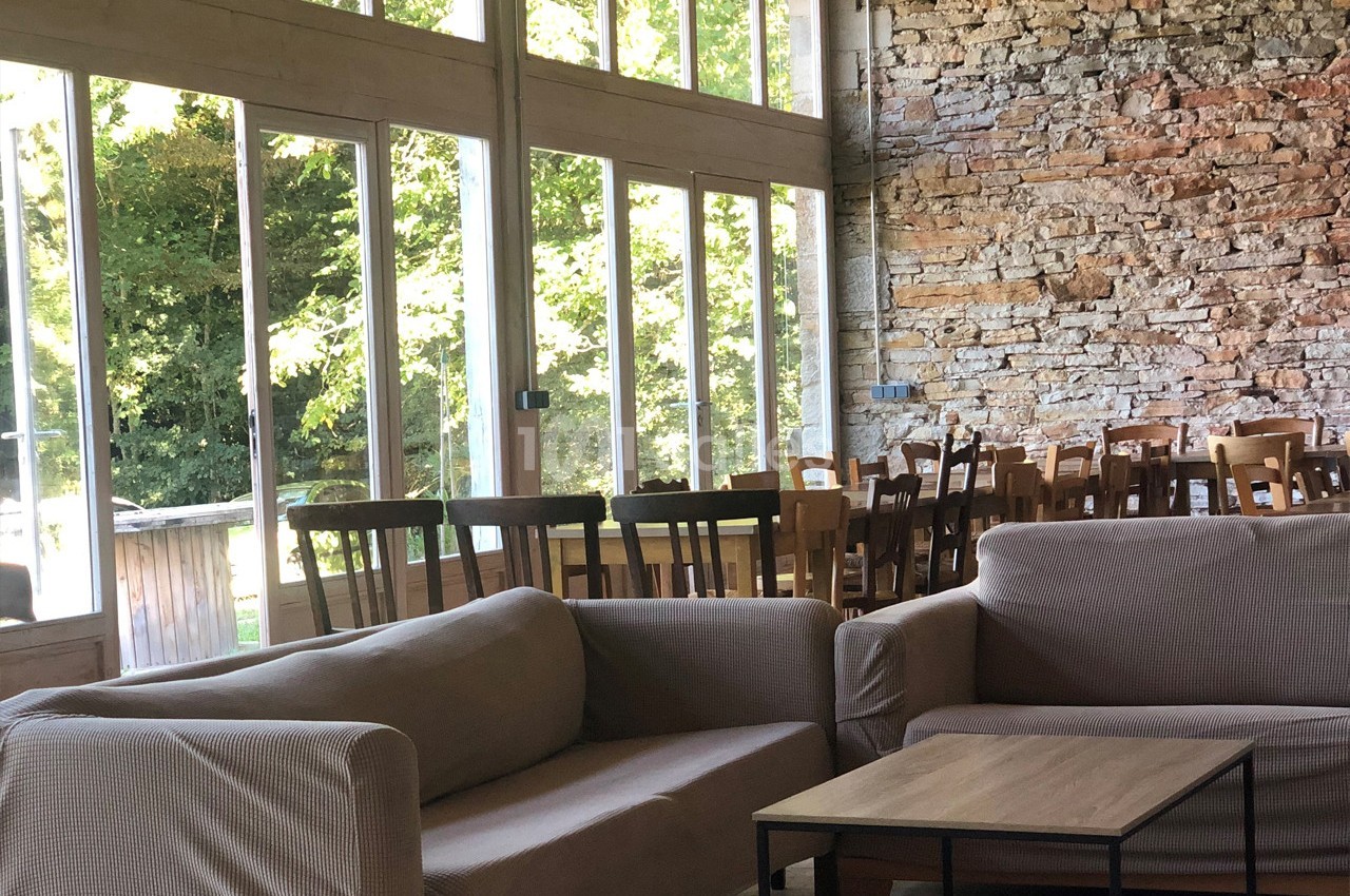 Salle lumineuse avec murs en pierre, grandes baies vitrées, canapés beiges et tables en bois alignées.