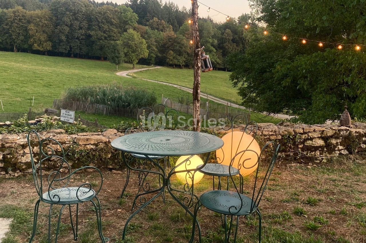 Table et chaises en fer forgé dans un jardin, éclairées par des guirlandes lumineuses et des lampes, avec vue sur un champ.