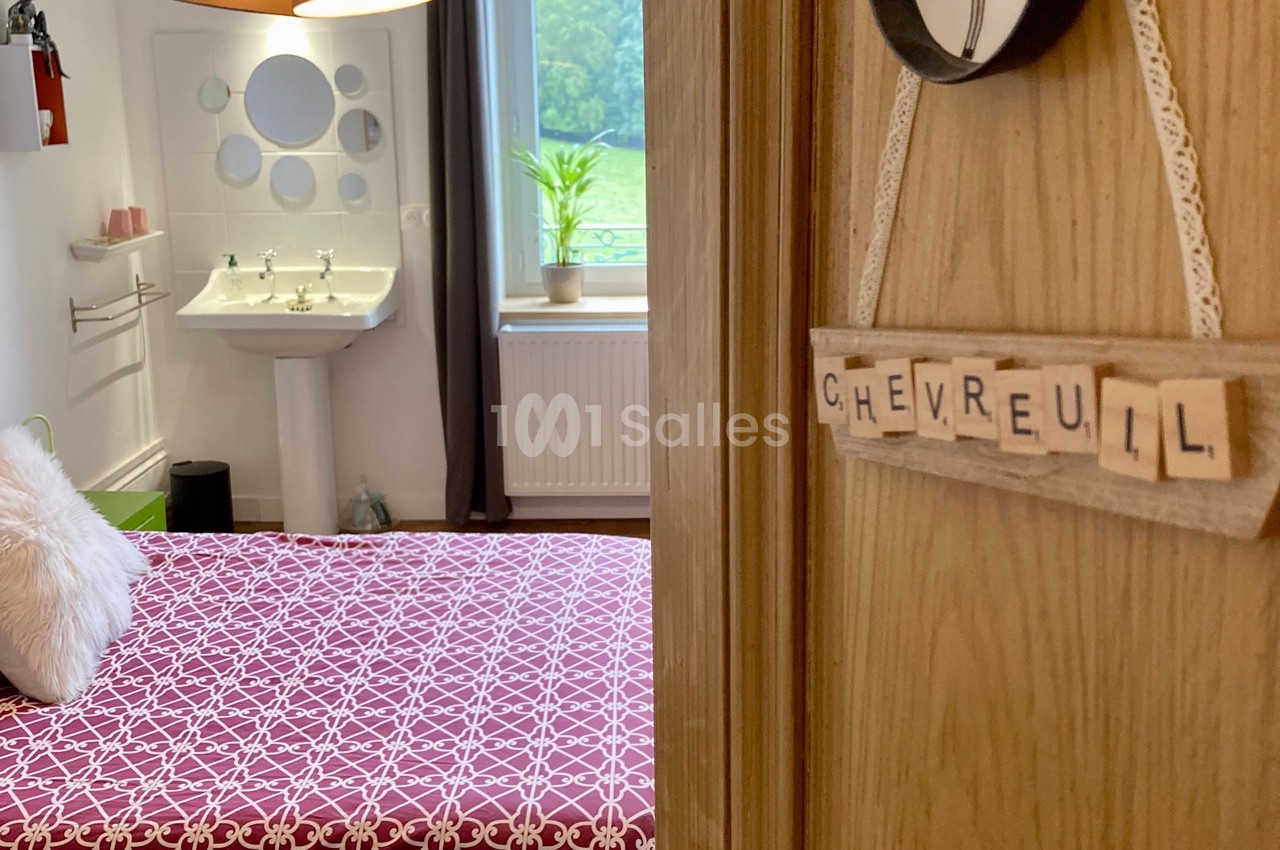 Vue d'une chambre avec un lit à motif géométrique, un lavabo, une fenêtre et une pancarte ’Chevreuil’ sur la porte.