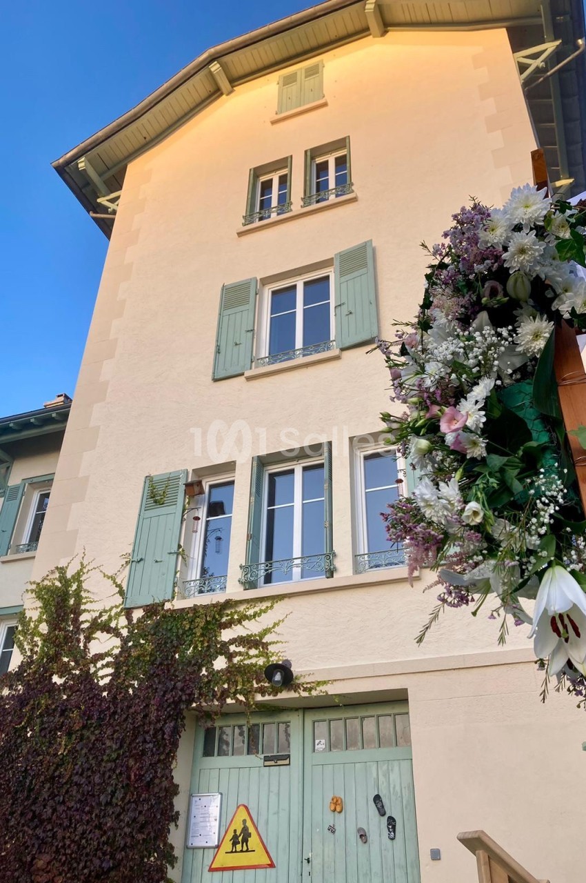 Façade d'une maison beige à volets verts, ornée de fleurs et de lierre, avec un panneau de signalisation devant.