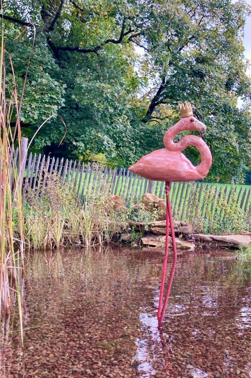 Statue d'un flamant rose avec une couronne, installée dans un étang peu profond entouré de végétation et d'une clôture en…