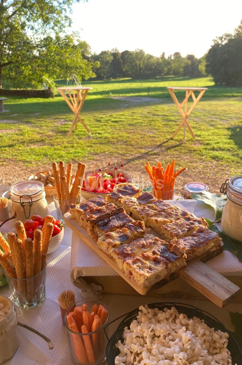 Table dressée en extérieur avec des plats variés, dont quiches, légumes croquants et popcorn, sur fond de prairie…