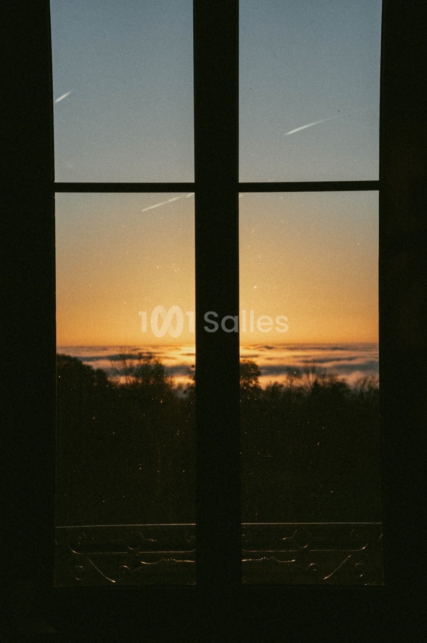 Vue d'un coucher de soleil à travers une fenêtre, avec des arbres en contre-jour et des traînées d'avion dans le ciel.