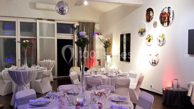 Salle de réception décorée avec des tables rondes dressées, des chaises blanches et des décorations murales colorées.