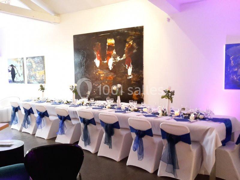 Table dressée pour un événement, ornée de nappes blanches, rubans bleus sur les chaises et décorations florales.