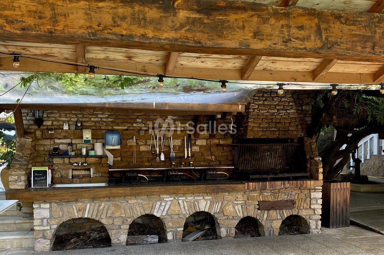 Cuisine extérieure en pierre avec un barbecue, des ustensiles suspendus et un toit en bois sous une pergola.