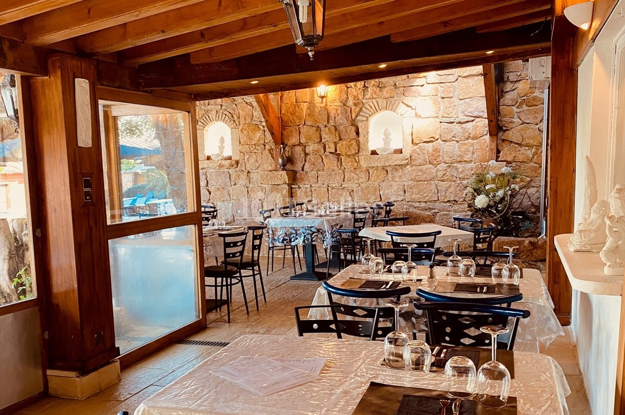Salle de restaurant lumineuse avec tables dressées, murs en pierre et poutres apparentes en bois.