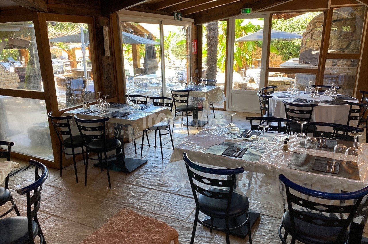 Salle de restaurant lumineuse avec tables dressées, chaises noires et vue sur une terrasse extérieure arborée.