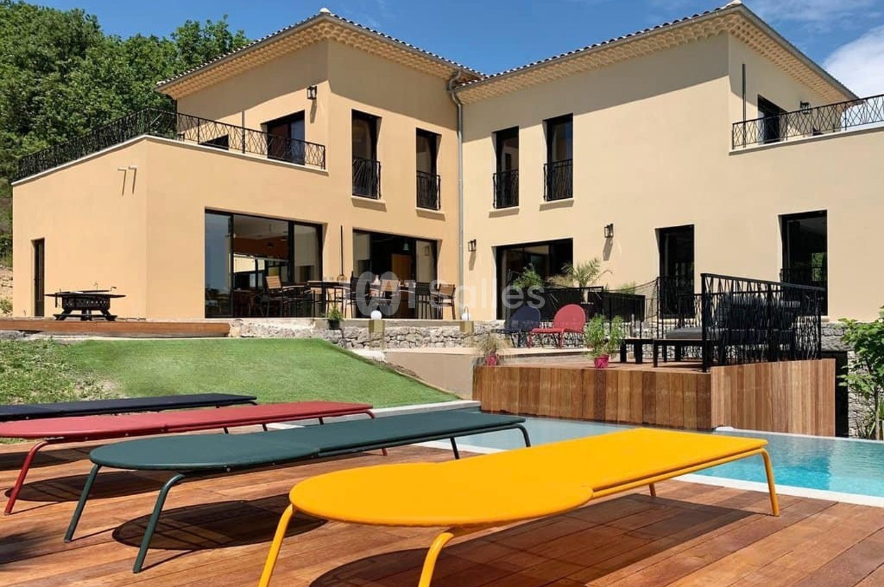 Façade d'une maison moderne avec terrasse, piscine et transats colorés sur un sol en bois, entourée de verdure.
