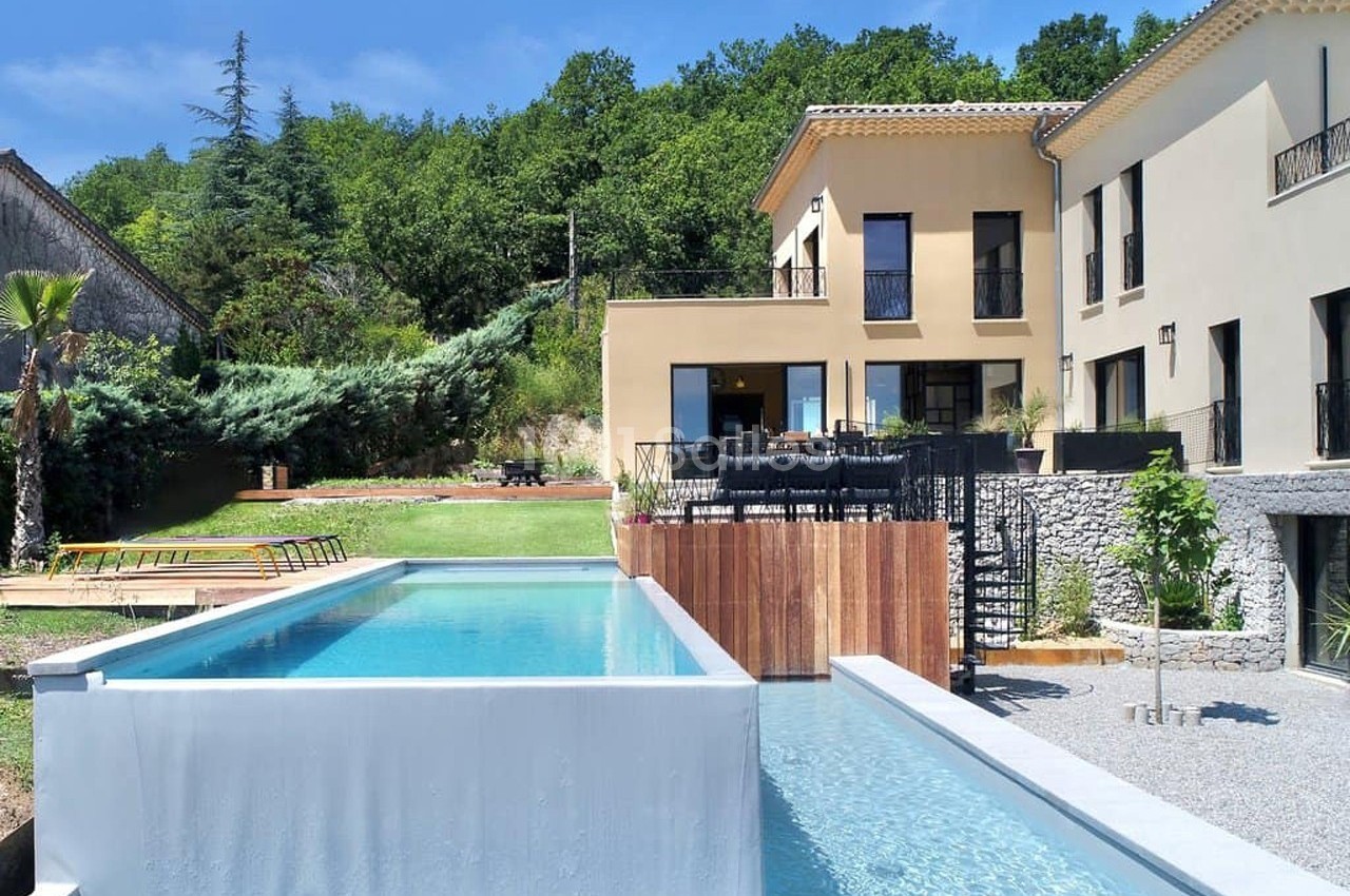 Maison moderne avec piscine à débordement, terrasse aménagée et jardin arboré, entourée de verdure.