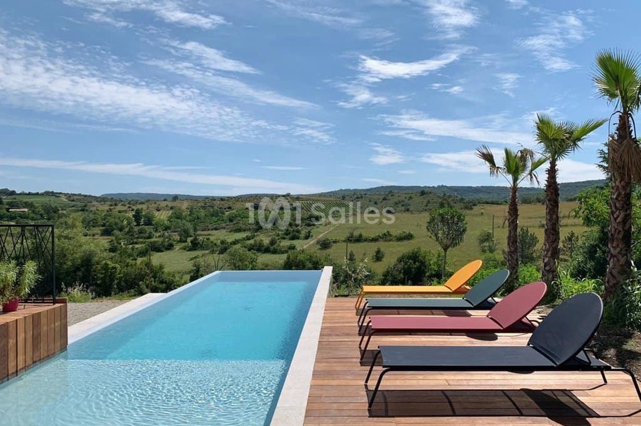 Piscine à débordement entourée de transats colorés sur une terrasse en bois, avec vue sur une campagne vallonnée.