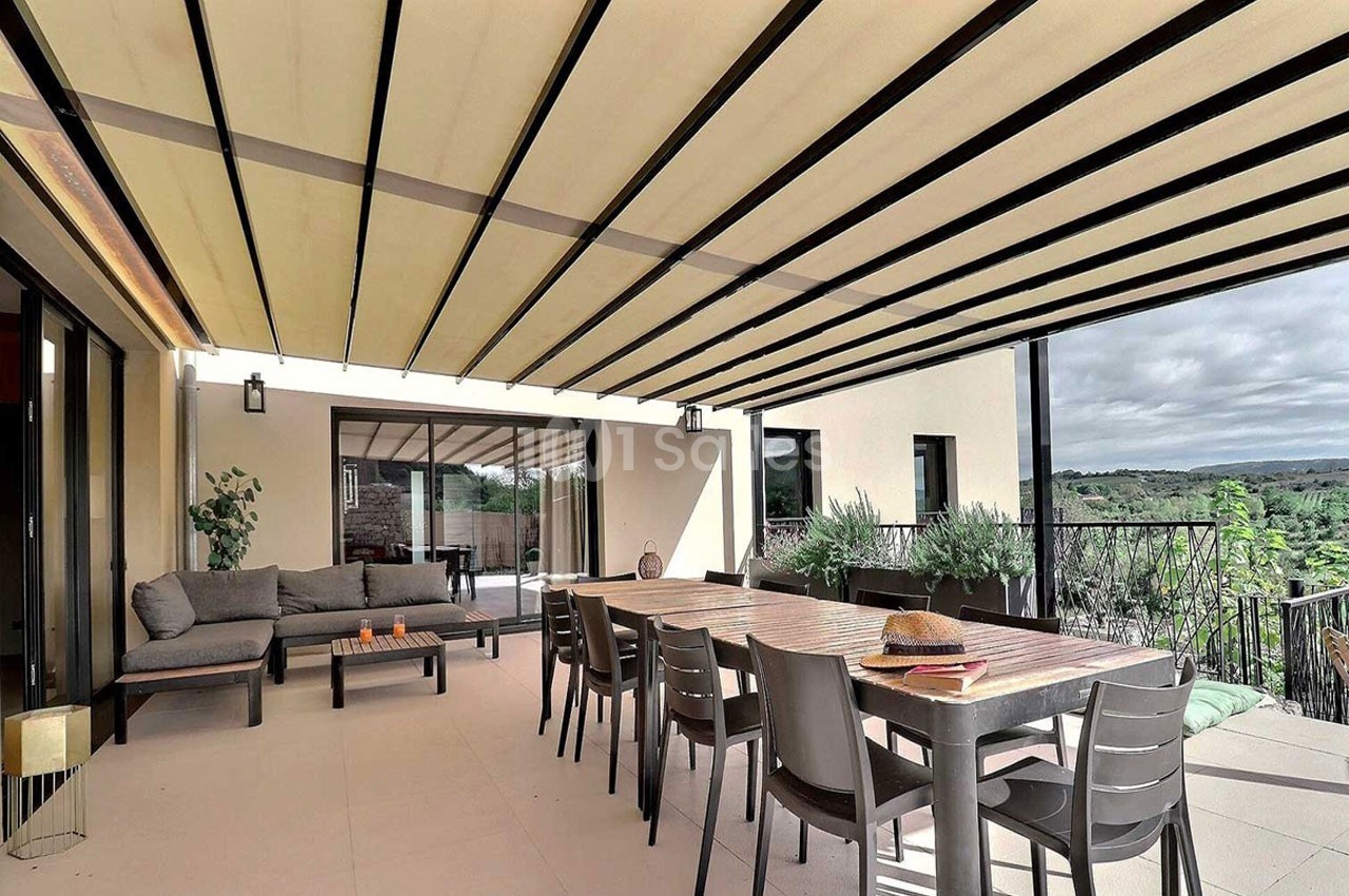 Terrasse spacieuse avec table, chaises, canapé et pergola à lames orientables, vue sur un paysage verdoyant.