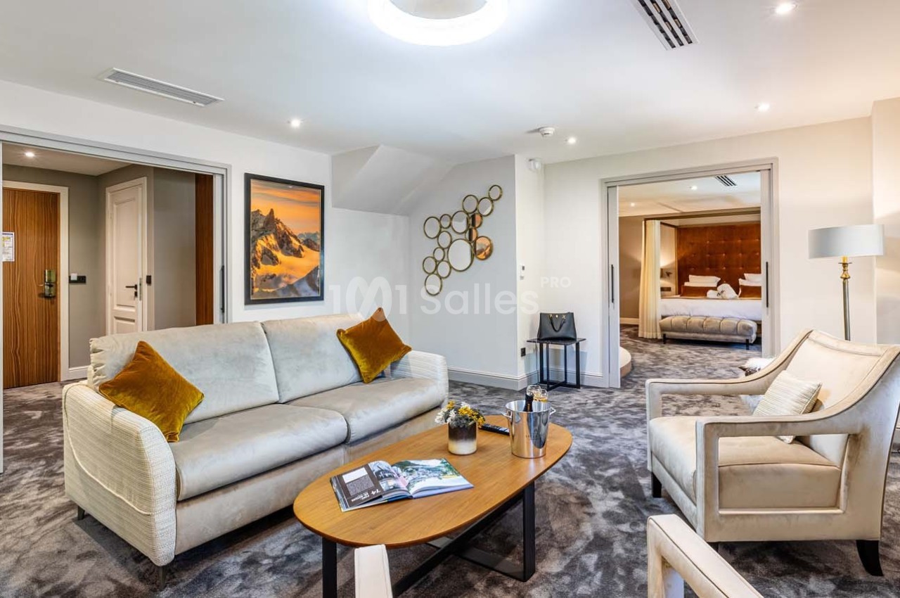 Salon d'un hôtel avec canapé, fauteuils, table basse et vue sur une chambre adjacente.