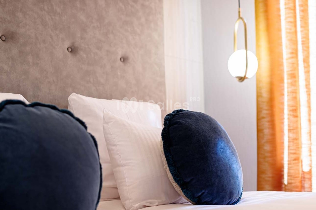 Lit avec coussins ronds bleu foncé, tête de lit capitonnée beige et rideaux orange laissant passer la lumière.