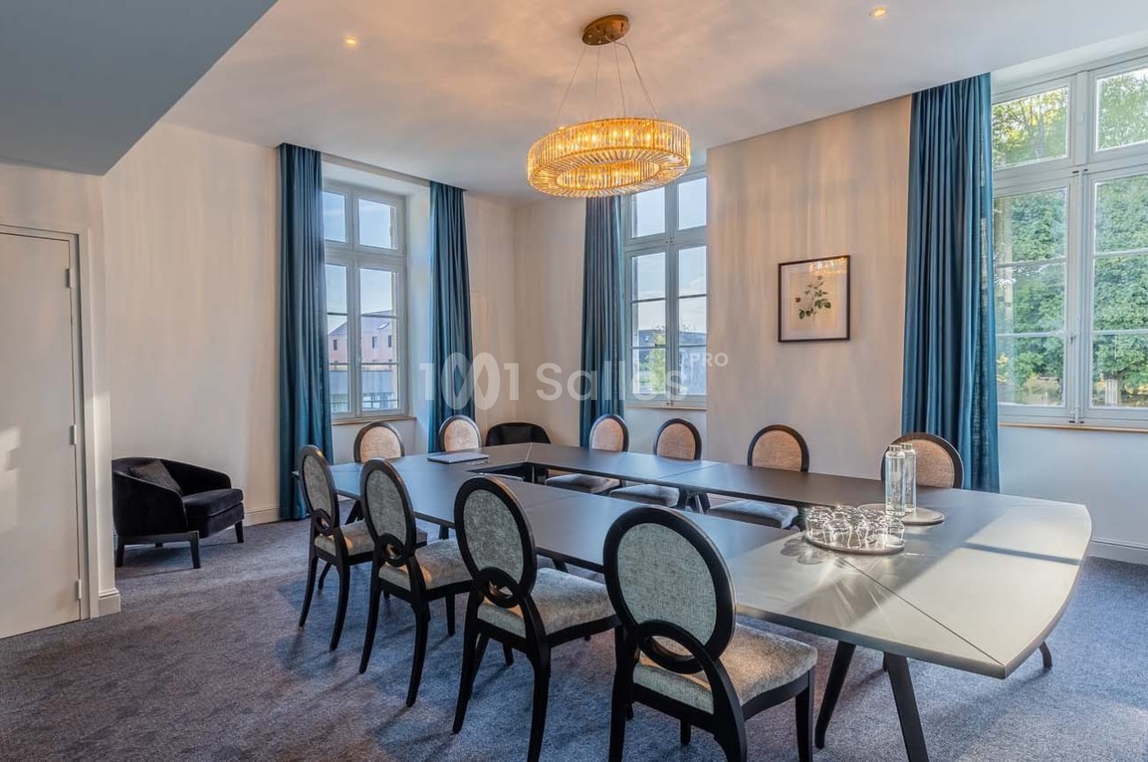 Salle de réunion lumineuse avec grande table ovale, chaises élégantes, lustre moderne et fenêtres donnant sur un extérieur…