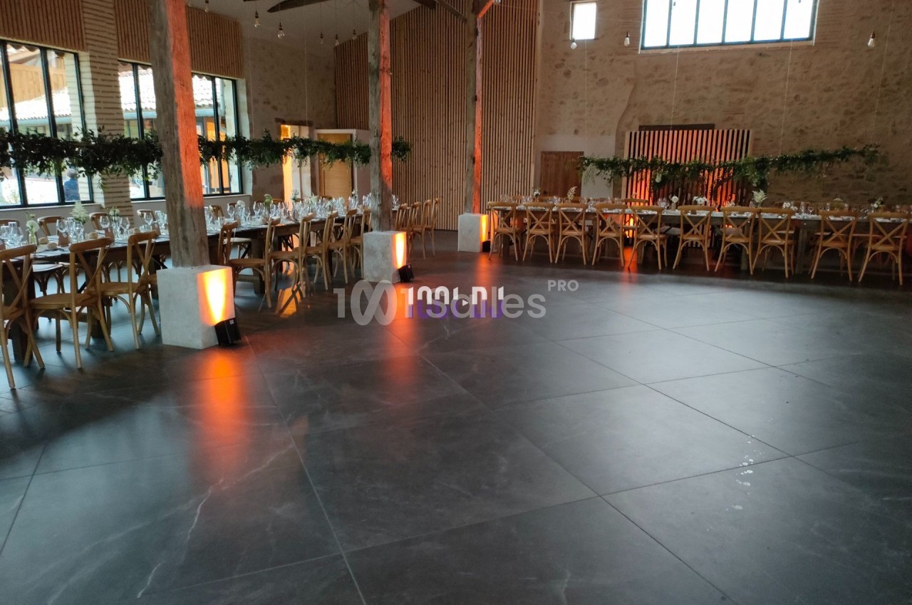 Salle de réception spacieuse avec tables dressées, chaises en bois et éclairage tamisé près des piliers.
