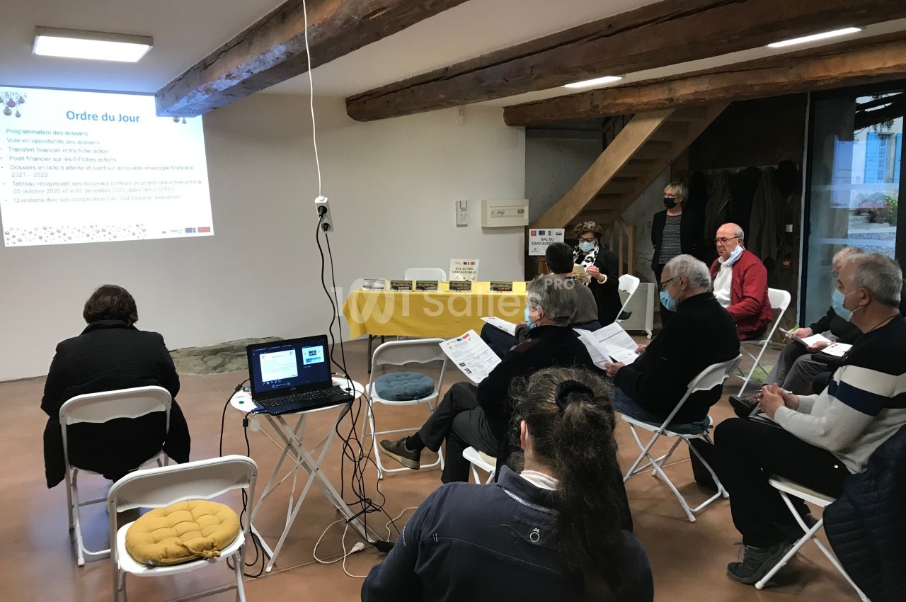 Des personnes assises dans une salle assistent à une présentation projetée sur un mur, avec documents en main.