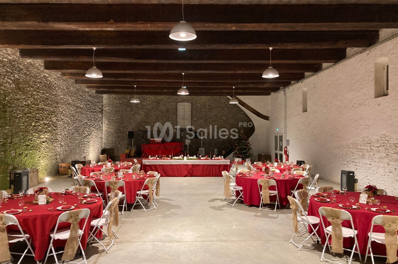 Salle de réception avec des tables rondes dressées de nappes rouges, chaises blanches et une scène au fond.