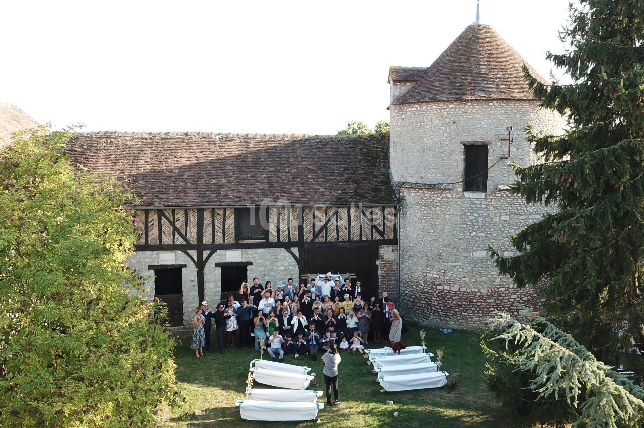 Groupe de personnes rassemblées devant un bâtiment en pierre avec une tour et un toit en tuiles, entouré de verdure.