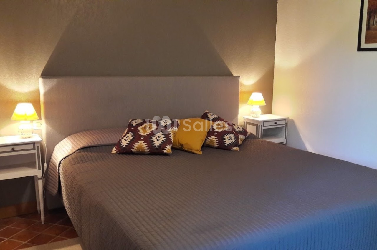 Lit double avec couvre-lit gris, coussins décoratifs colorés, deux lampes de chevet allumées et mur beige en arrière-plan.
