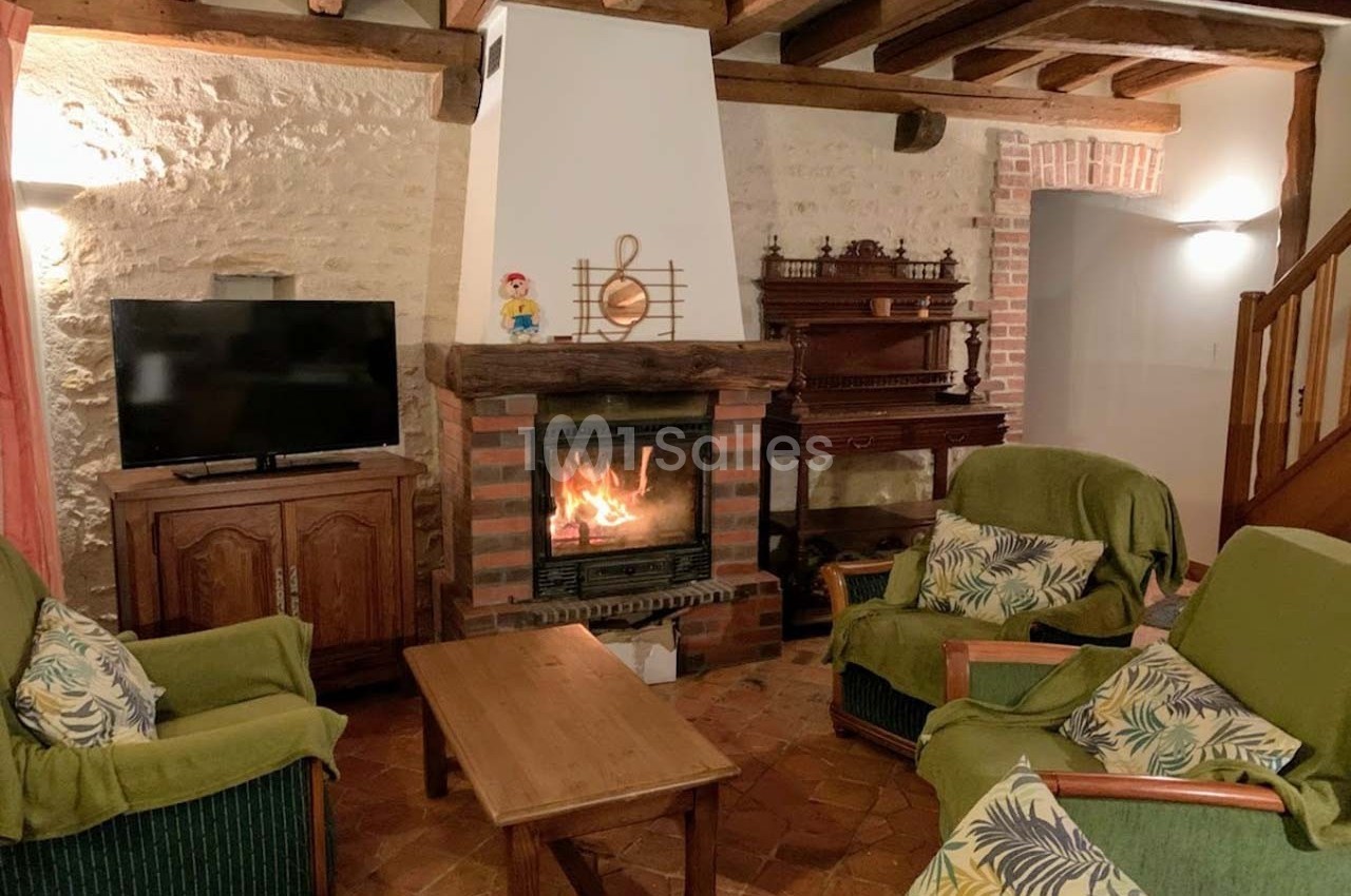 Salon chaleureux avec cheminée allumée, meubles en bois, fauteuils verts et murs en pierre apparente.