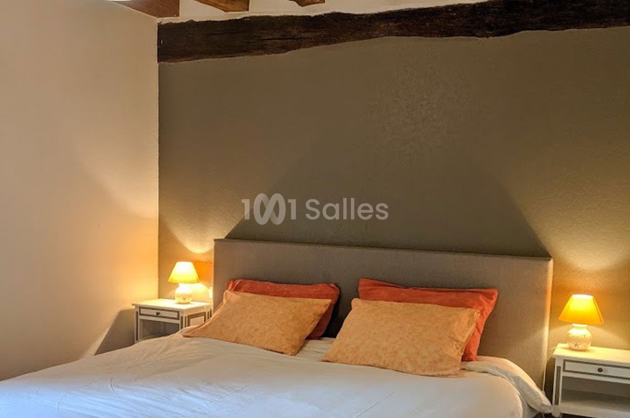 Lit double avec tête de lit grise, deux tables de chevet blanches et lampes allumées dans une chambre aux poutres apparentes.