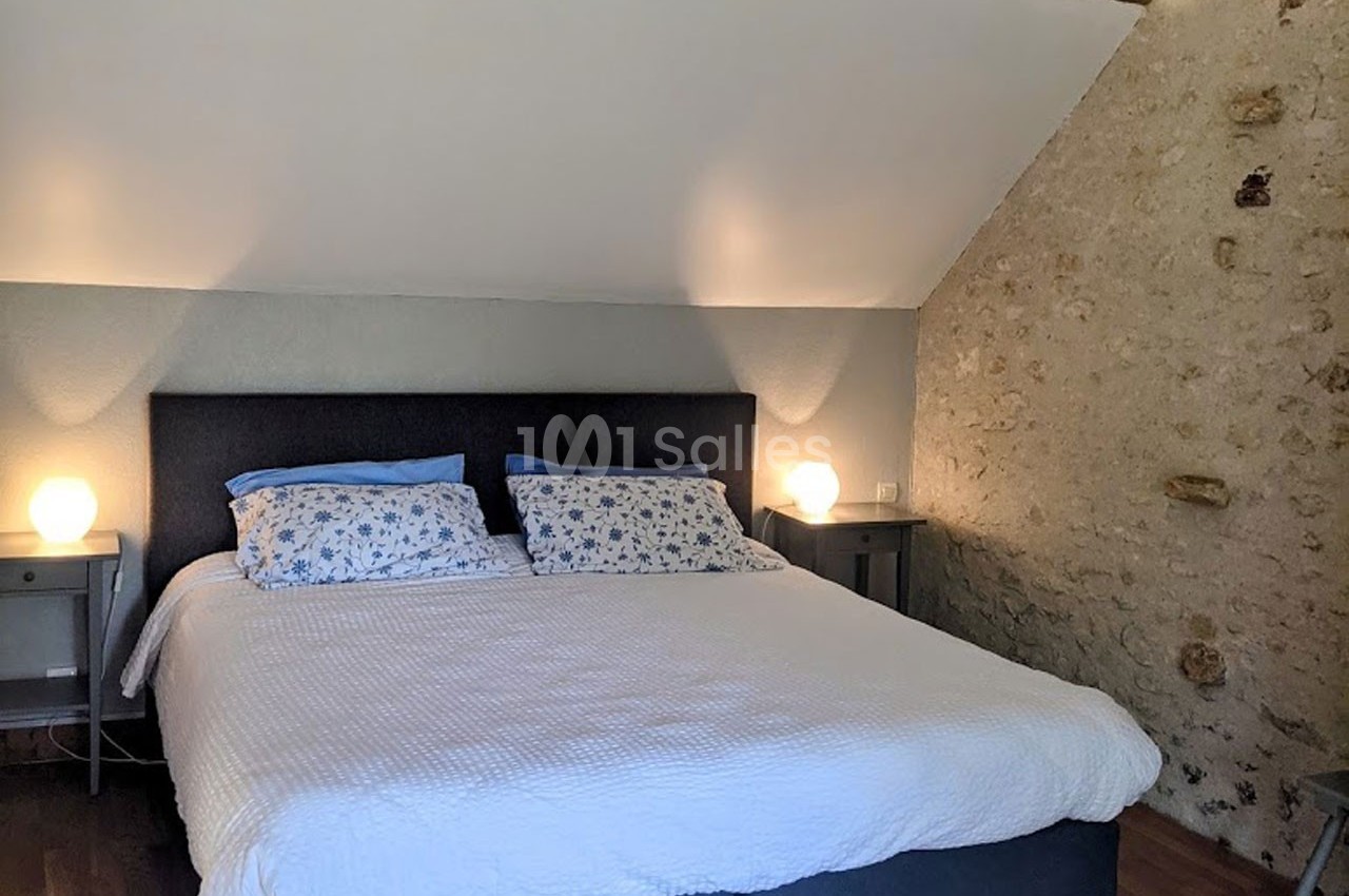 Lit double avec tête de lit grise, draps blancs et coussins à motifs, dans une chambre aux murs en pierre et parquet.