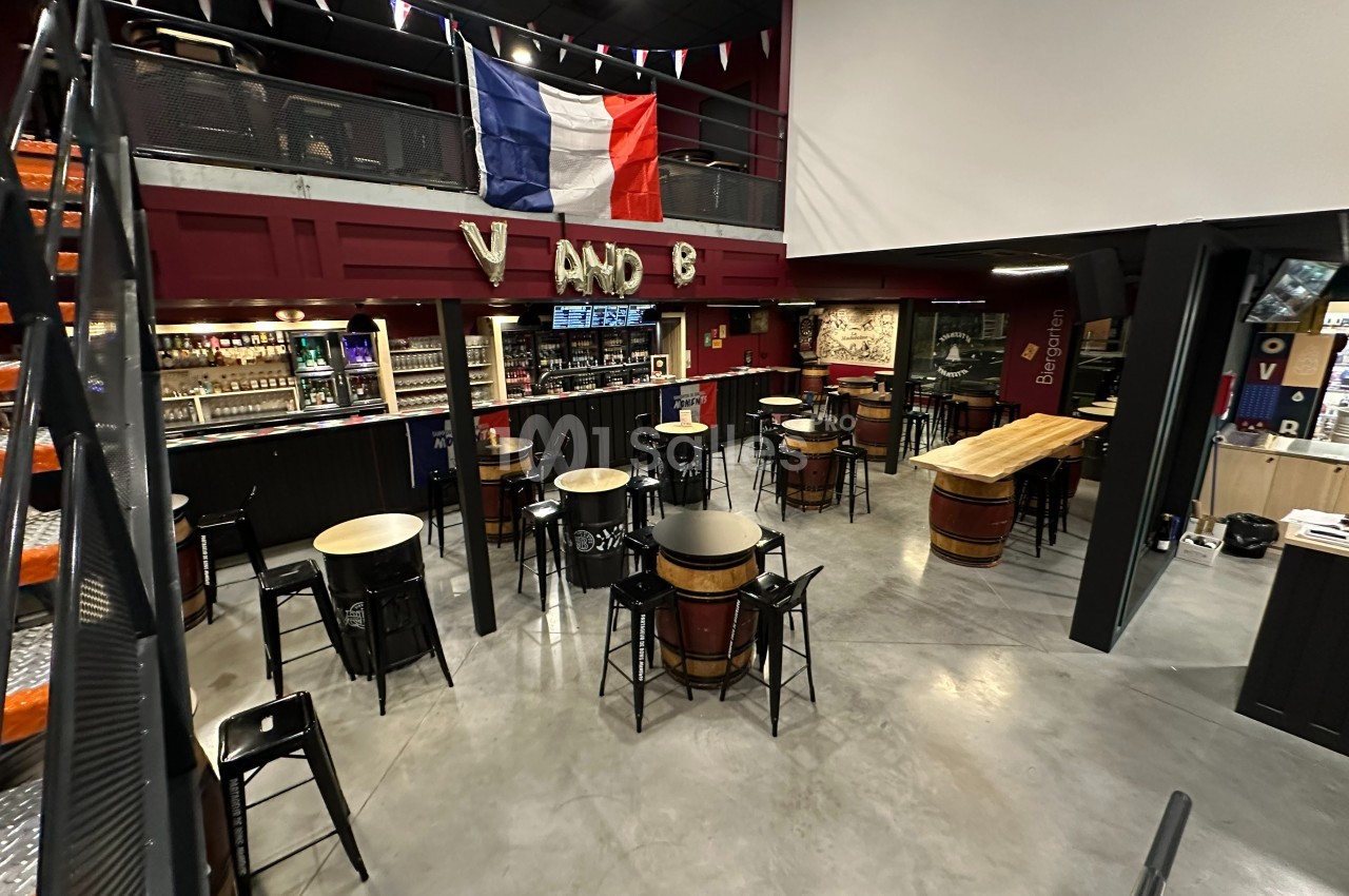 Intérieur d'un bar avec tables rondes, tabourets hauts, comptoir en bois et drapeau français suspendu.