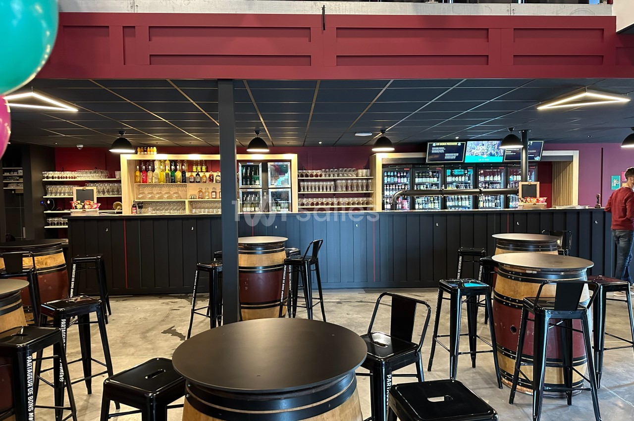Salle de bar avec tables en tonneaux, chaises hautes, comptoir en bois et étagères garnies de bouteilles.
