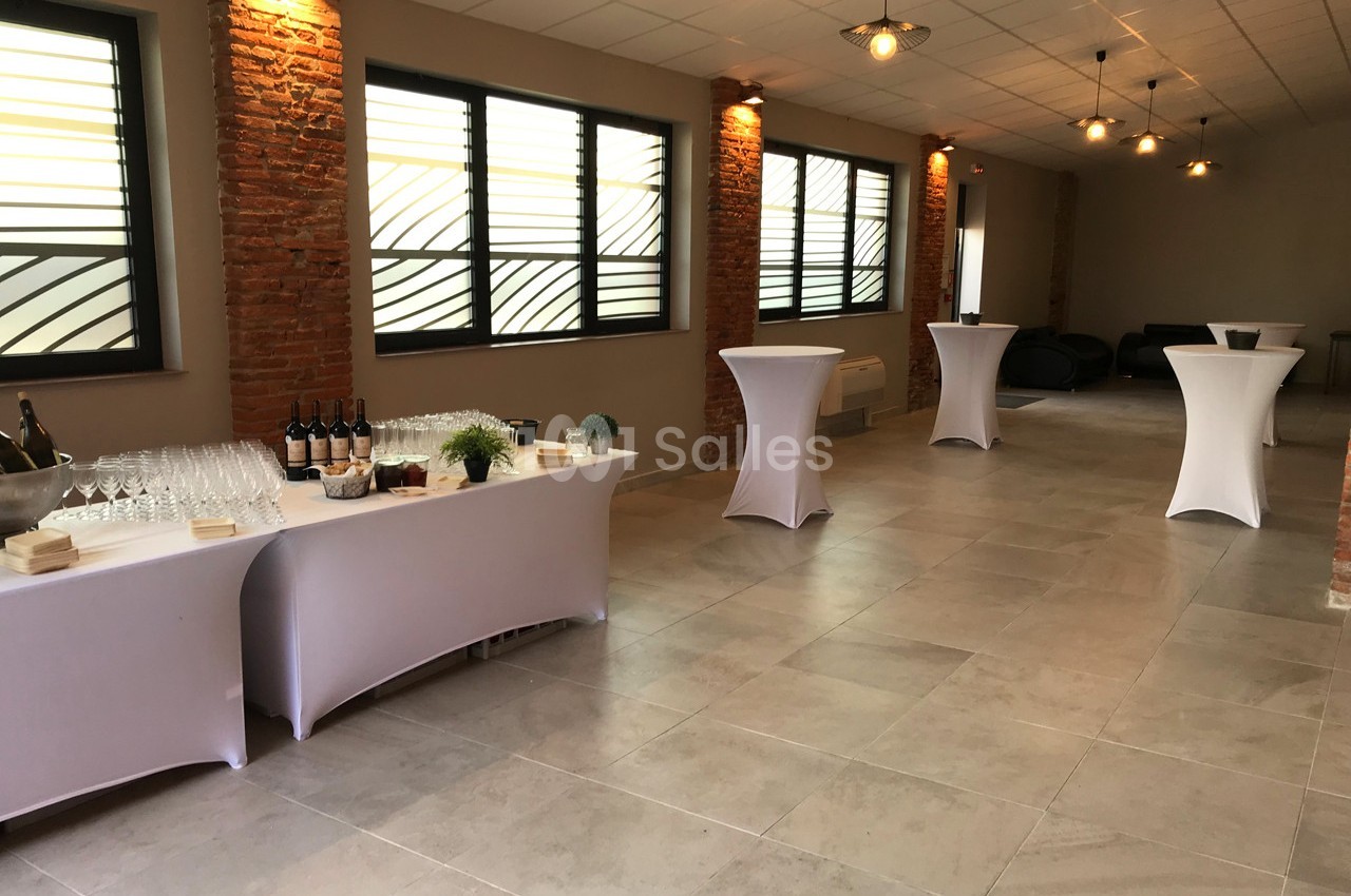 Salle lumineuse avec tables hautes drapées de blanc, buffet de boissons et verrerie, murs en briques et grandes fenêtres.