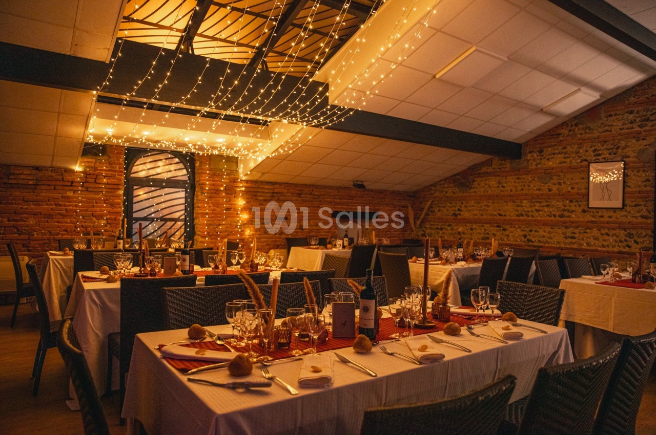 Salle de réception décorée avec des guirlandes lumineuses, tables dressées avec nappes blanches et vaisselle élégante.