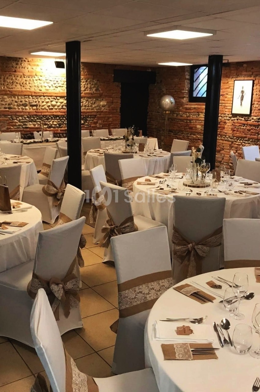 Salle de réception avec tables rondes dressées, nappes blanches et décorations en rubans beige et dentelle.