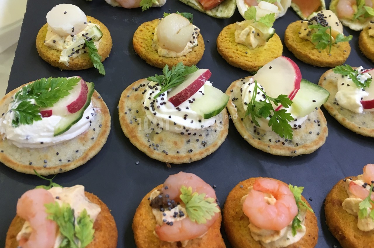 Assortiment de canapés garnis de légumes, crevettes, fromage frais et herbes, présentés sur un plateau noir.