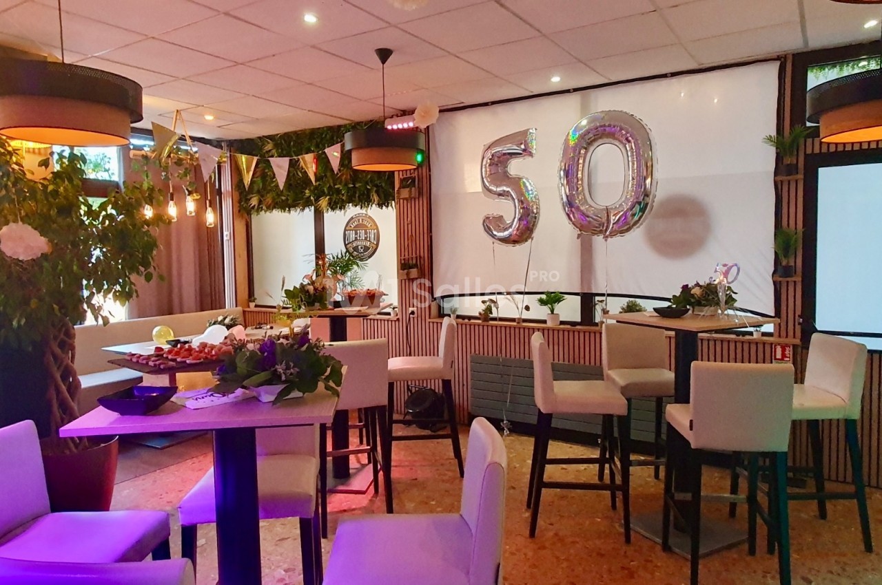 Salle décorée pour une fête avec des ballons ’50’, des guirlandes, des plantes et des tables hautes entourées de chaises.