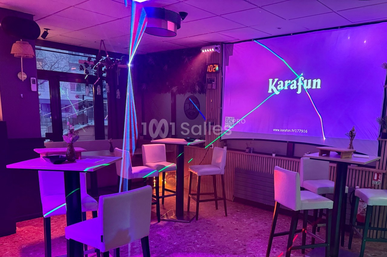 Salle de karaoké avec des tables hautes, des chaises blanches et un écran affichant ’Karafun’ sous des lumières colorées.