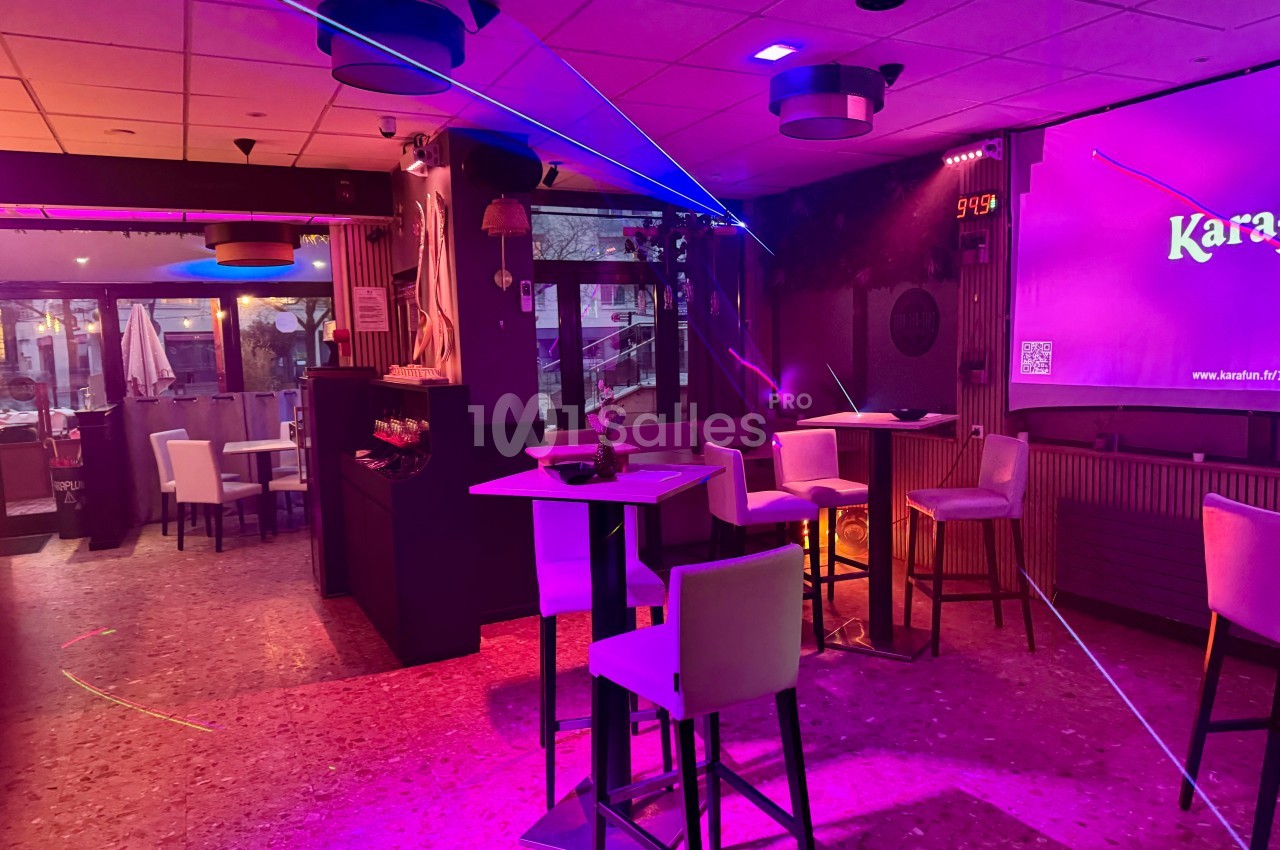 Salle de karaoké avec éclairage coloré, tables hautes, chaises blanches et vue sur une terrasse extérieure.