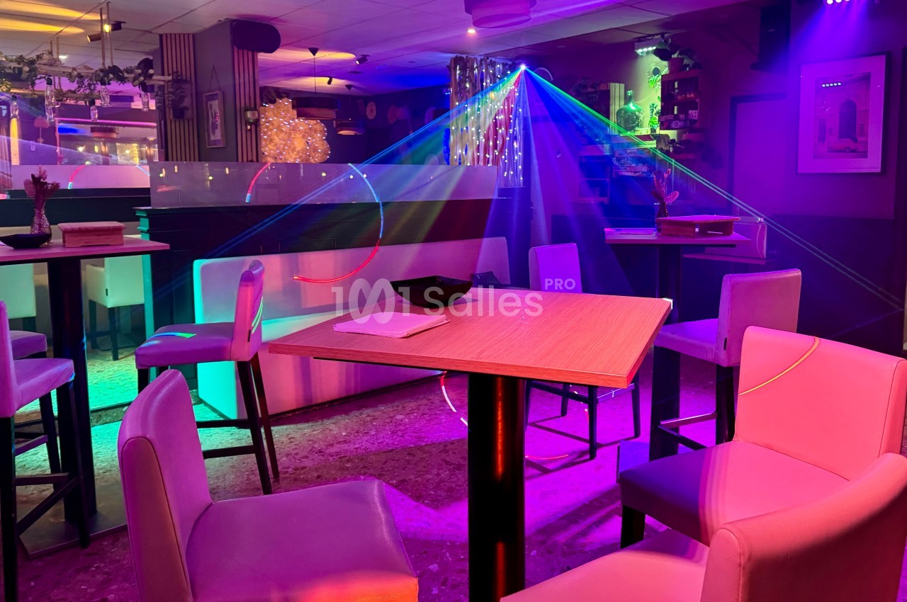 Salle de bar ou restaurant avec éclairage coloré, tables et chaises modernes, et ambiance festive.