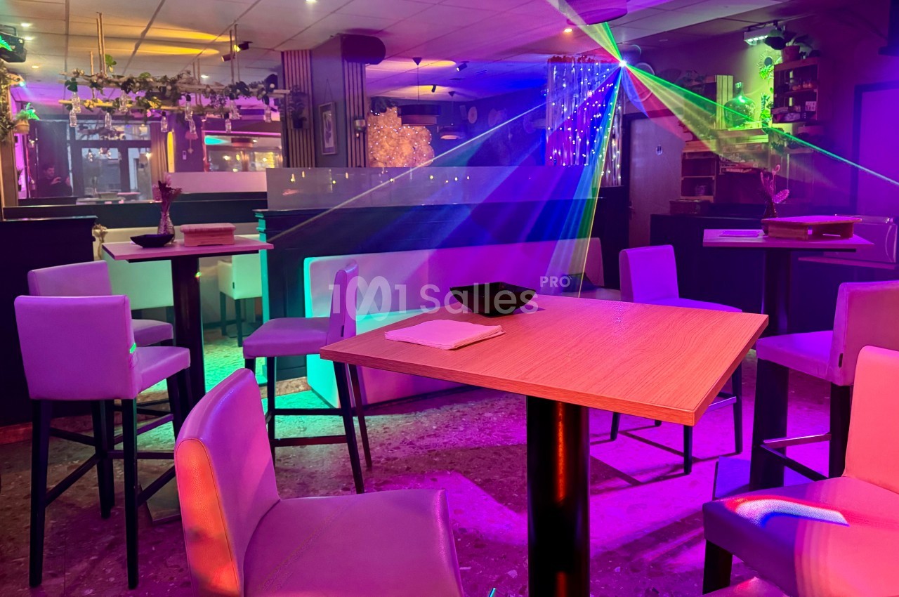 Salle de bar avec tables hautes, chaises, éclairage coloré et jeux de lumière dans une ambiance nocturne.